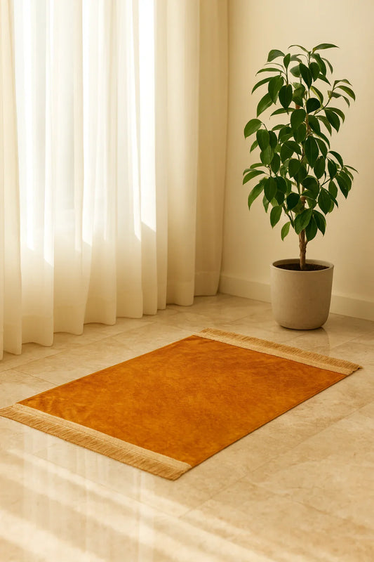 Simple orange Islamic prayer mat with fringe on polished floor near potted green plant and white curtains
