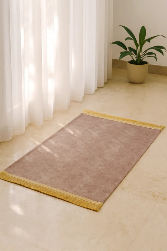 soft brown Islamic prayer mat with golden fringes on marble floor near sheer white curtains and green potted plant